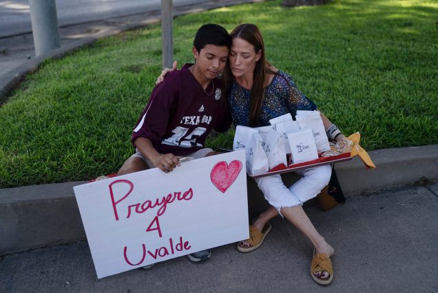 Photo of Diego Esquivel, left, and Linda Klaasson sit on curb at the edge of a green space and comfort each other as they gather to honor the victims killed in Tuesday's shooting at Robb. Esquivel is a young, Latinx tween boy who wears a crimson Texas jersey. He holds a sign that says "Prayers 4 Uvalde." Klaasson is a middle aged woman with light-brown skin and long brown hair. She closes her eyes as she gives Esquivel a side a hug. Balanced on her lap is a tray of white paper bags with presumably food in them; blue label on the bag reads "the Core" and has a logo of an apple core with a couple bites out of it.