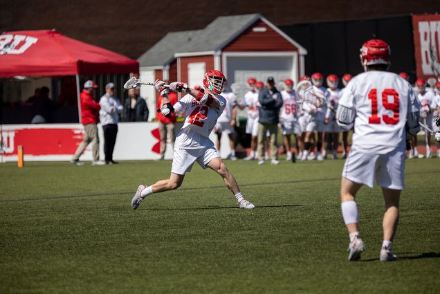 Photo of BU men's lacrosse player Jake Cates. Action shot shows him mid throw, lacrosse stick up as he wears a white BU lacrosse uniform and red helmet. Similarly dressed players can be seen standing on the sidelines of the field.