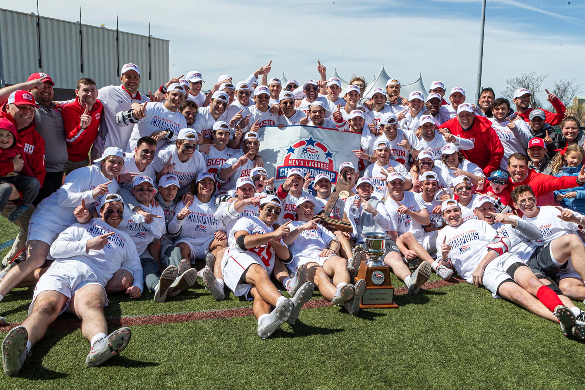 Group photo of the moments after BU claimed its first conference trophy. The mens lacrosses players sit and stand in white in a group with their coaches in red, and various family members. Everyone wears white hats and in the middle of the group they hold a sign that reads "Patriot League Men's Lacrosse Championship". a large silver trophy sits on the ground in front of the group.