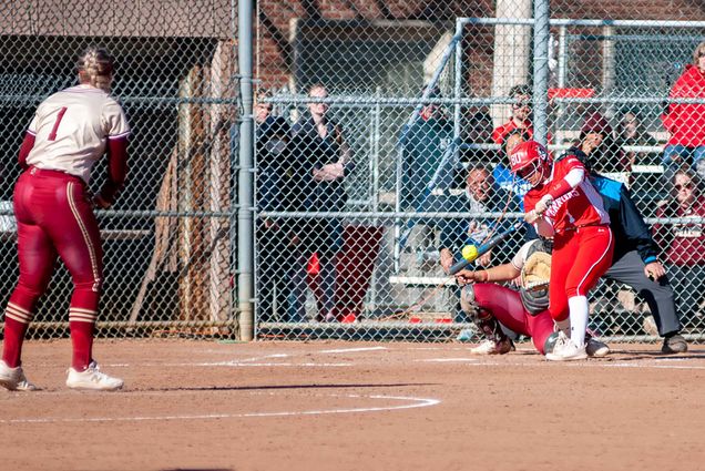 Action photo of Marina Sylvestri. She is caught mid-swing, about to hit a softball thrown by the opposing pitcher wearing a burgundy uniform. Spectators can be seen on the other side of the fence behind homebase, watching on.