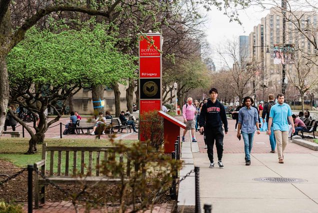 Photo of a diverse group of students walking down Comm Ave on a spring day. A red BU sign is seen at left, as well as a tree with budding green leaves.