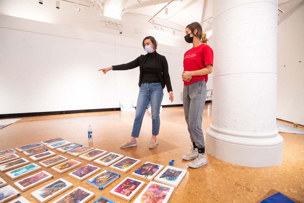 Photo of Leah Triplett Harrington, guest curator of this year’s BFA Thesis Art Exhibit,  advising painting major Anna Frants (CFA’22) during installation of Frants’ work. Both wear face masks and stand looking at a series of paintings on the ground in front of them; each canvas is about 8x11 inches and features abstract portraits. 