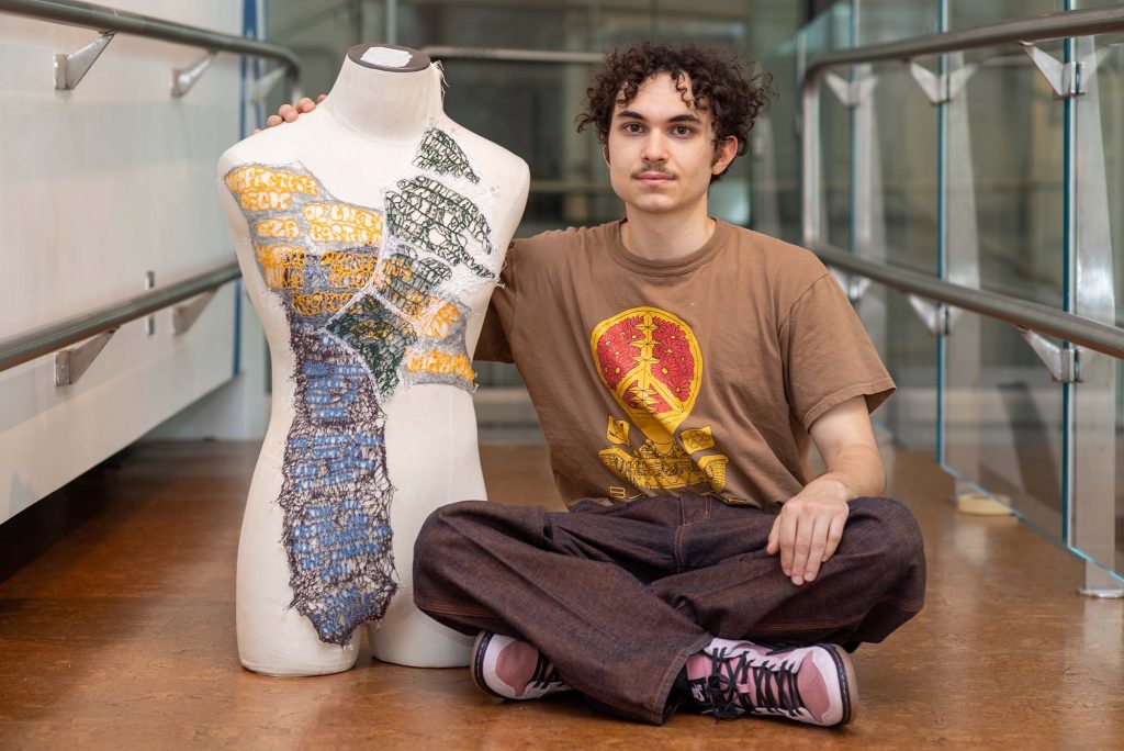 Photo of Jonathan Pinchera (CFA’23) sitting cross-criss next to his piece Narrative Lace at the BFA Thesis Art Exhibit at Stone Gallery May 5. Pinchera has short curly hair and a mustache, and wears a brown t-shirt and dark brown pants. The lace artwork is stitched onto a mannequin's torso; yellow, blue and green bubble letters are seen inside the design.