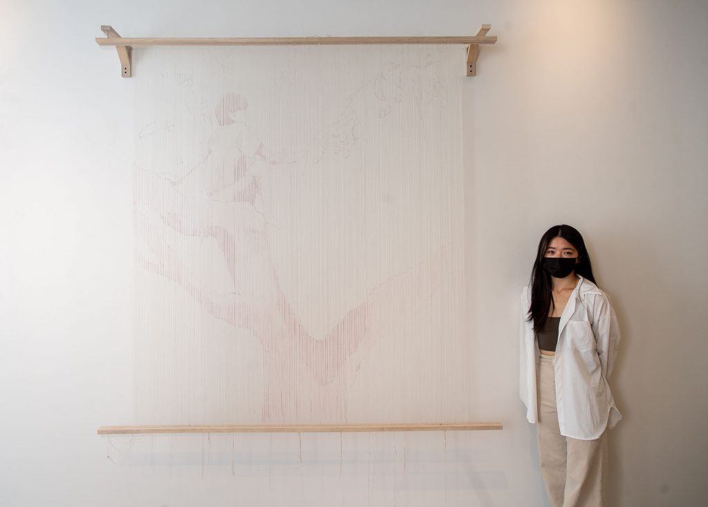 Photo of Ashley Zhou (CFA’22), a young AAPI student who wears a black face mask, tan pants, and a flowing white shirt, standing to the right of her piece called Cherry Tree at the BFA Thesis Art Exhibit at Stone Gallery May 6. The piece is a tapestry and a design of a tree with what looks like a woman sitting in it is made barely visible by light red stands of thread.