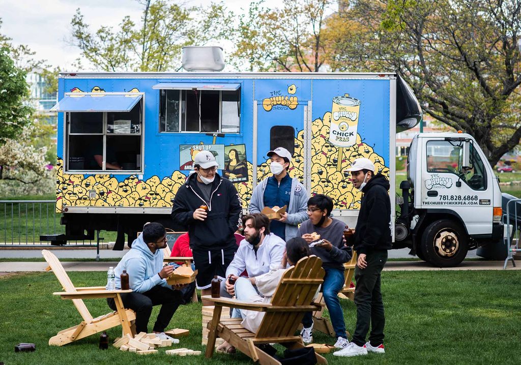 Photo of Students enjoying themselves on BU Beach during Senior Week. A few students stand while others sit as they east and chat on a grassy lawn. A blue food truck can be seen behind them.