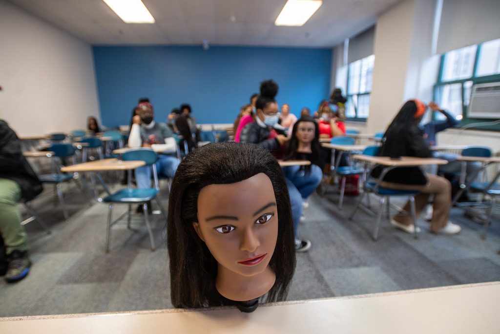Photo of the Naturally BU club preparing for their braiding demonstration in CAS. Students enter the room and sit at desks in the blurry background. In the foreground, a mannequin with thick, kinky straight hair, is clipped to a table in the foreground.