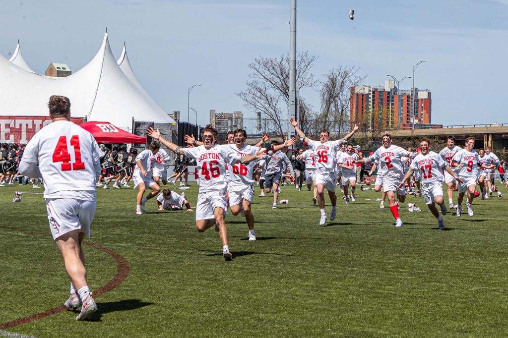 Photo of the lacrosse Terriers running to celebrate with Patriot League Goaltender of the Year Matt Garber. They all wear white uniforms as they run with arms int he air, cheering.