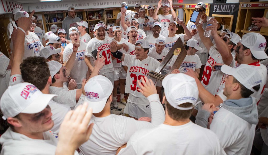 Photo of Cocaptain Matt Hilburn holding up the Patriot LEague trophy among the cheering men's lacrosse team in their locker room. All wear white uniforms and white caps as they cheer and shout in joy.