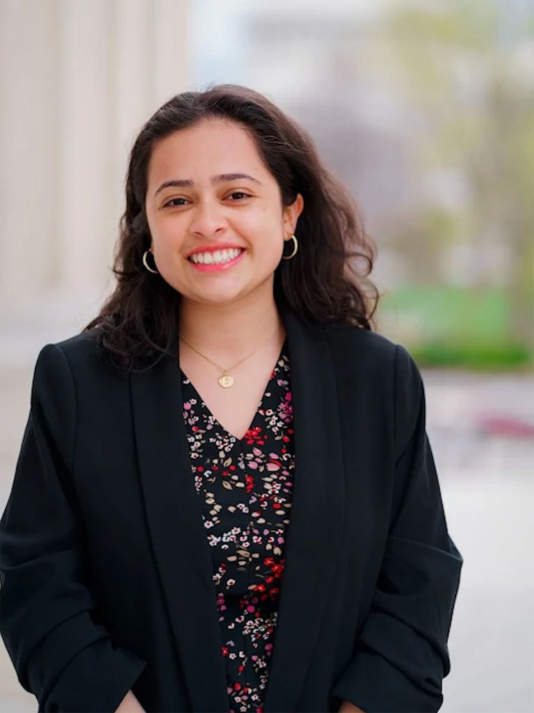 Samara Parada smiles for the camera in a suit jacket, wearing earrings and a necklace as well as a top with a flower pattern. The background is blurred