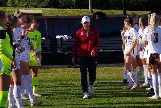 Photo of Casey Brown walking down a soccer field between two rows of Penn players in white. She wears a white cap, red pants with a Penn logo, and black pants.