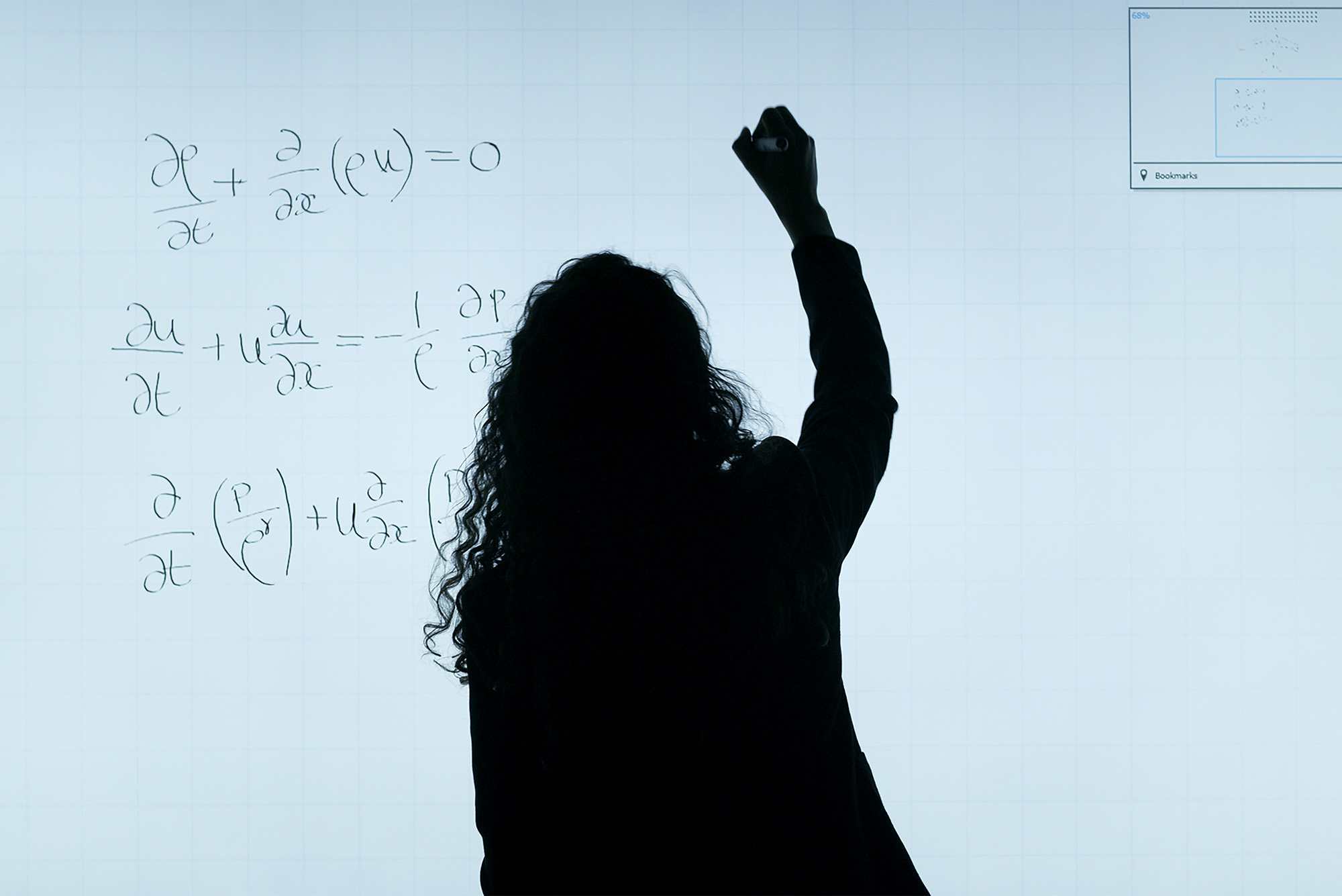 Photo of a silhouette of a person with long curly hair writing equations on a clear board