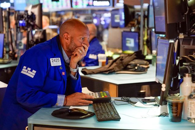 Photo of a trader (a White, bald man wearing a blue jacket) working on the floor at the New York Stock Exchange in New York, Tuesday, June 14, 2022. He has his left hand on his face and looks concerns as he types at a computer screen. Other people working on computers and large screens with stock information on them are seen blurred behind him.