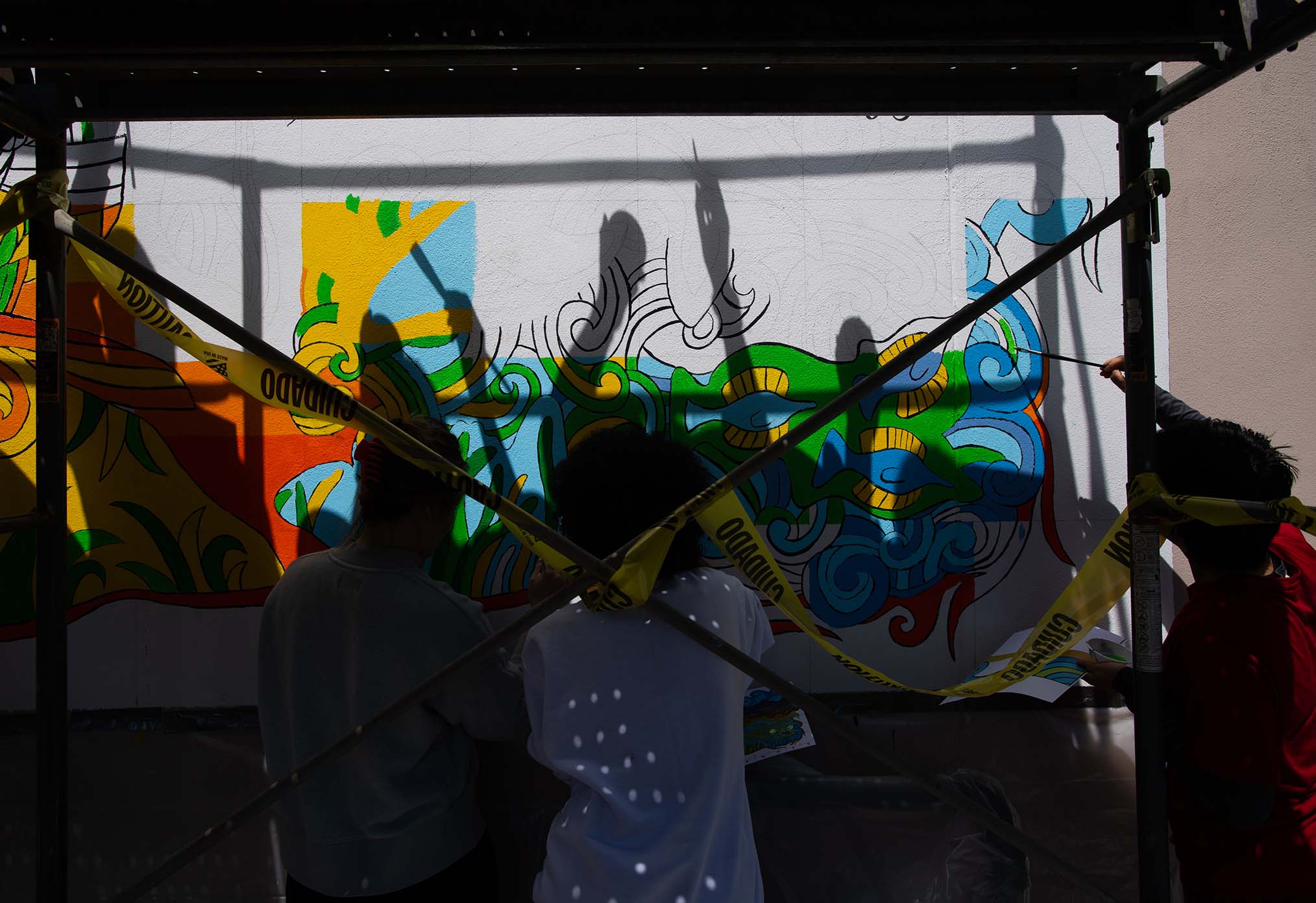 Photo of three students working together on a collaborative mural on the side of a white building. Parts of the mural are painted in with bright colors and students work under the shadow of a large awning. 