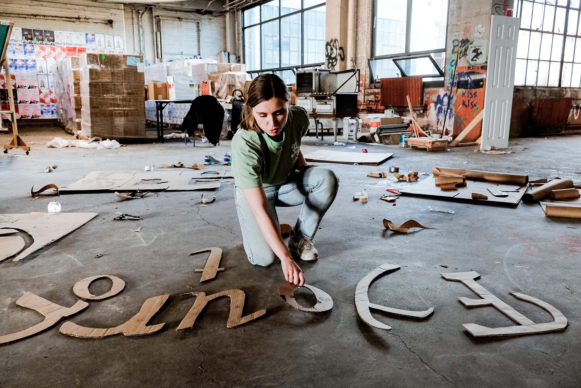 Photo of Mackenzie Bower checking the size and font of a finished letter template. A young white woman with brown hair and wearing a light blue shirt and jeans arranges large, tan cutout letters on the dark cement ground of a bright warehouse.