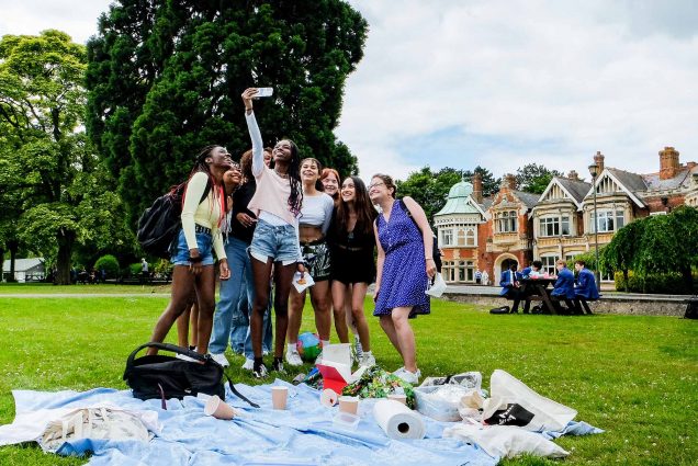 Photo of a diverse of students, most of them women, taking a selfie behind a large blue picnic blanket. The women smile as a student of color holds up her phone to take the selfie. The grass of the park is bright green and British houses and a large tree can be seen in the background.
