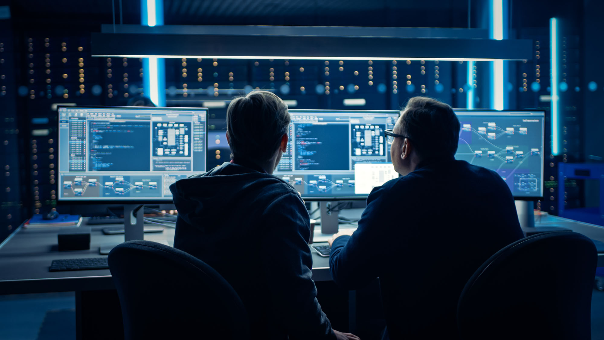 Photo of two people sitting in front many screens filled with code and information. Screens are toned a bright, neon blue as the backs of the two people can be seen in shadow.