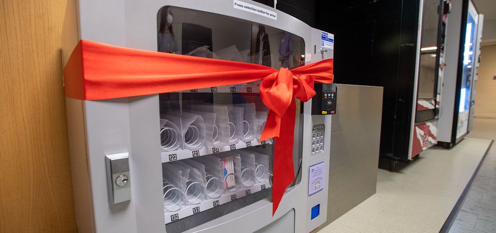 Photo of a small grey vending machine. Most of the vending slots are empty in the photo, except for a few that have plan b in them. A red ribbon is tied around the machine.