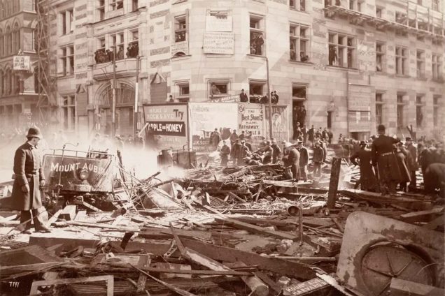 Sepia photo of the aftermath of a devastating gas line explosion at the corner of Tremont and Boylston streets on on March 4, 1897, which blew up two trolley cars and left 10 people dead, just six months before the Boston subway was scheduled to open. At left, a man in a bowl hat and a long trench coat looks out into the rubble of metal and piece. A crowd of people, back right, can also be seen examining the damage. Photo by Nathaniel Livermore Stebbins, courtesy Boston Pictorial Archive via Boston Public Library