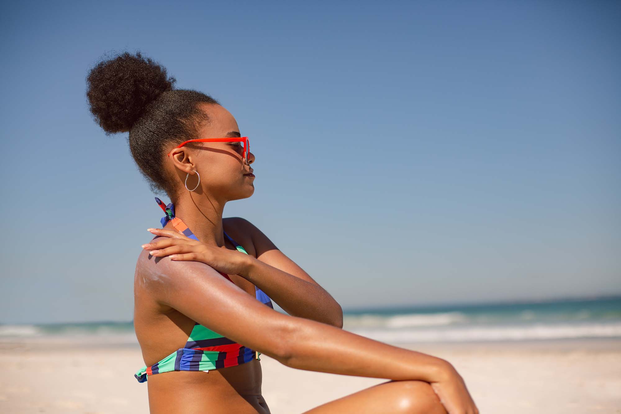 Photo taken from the side of a Black woman in a colorful, striped bikini applying sunscreen lotion on her right shoulder at beach in the sunshine. She sits on the ground and looks towards the water as she applies sun screen. She wears red sunglasses and silver hoop earrings. A clear blue sky and small waves crashing can be seen blurred in the background.