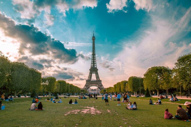 Photo of the Eifel tower seen from a grassy park lined with manicured trees on a summer night with a circle of clouds around the towers. Small groups of people enjoy the evening in the park.