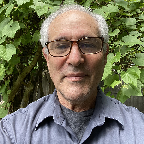 Photo of Richard Reibstein. An older man with grey hair and glasses wearing a blue collared shirt looks into the camera and stands in front of greenery.