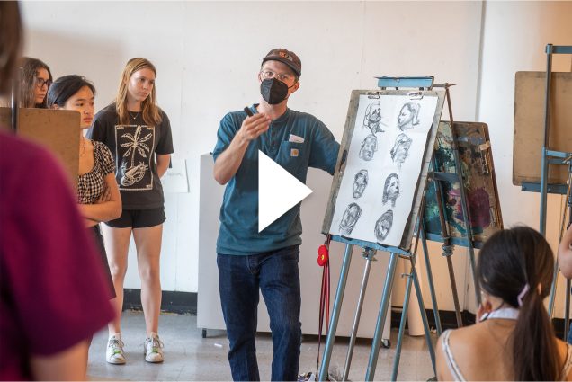 Photo of Instructor Sam Guy with his VASI drawing class. A masked man wearing a blue VASI labeled shirt and black face mask speaks to students as he stands in front of a metal easel featuring various charcoal sketches of heads. He holds a stick of charcoal in his hand as he gesticulates. A white, triangle play button is overlaid on the image.