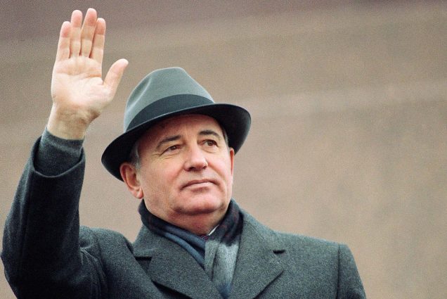Photo of Gorbachev waving. An older white man wearing a black wool coat and hat waves to an unseen crowd.