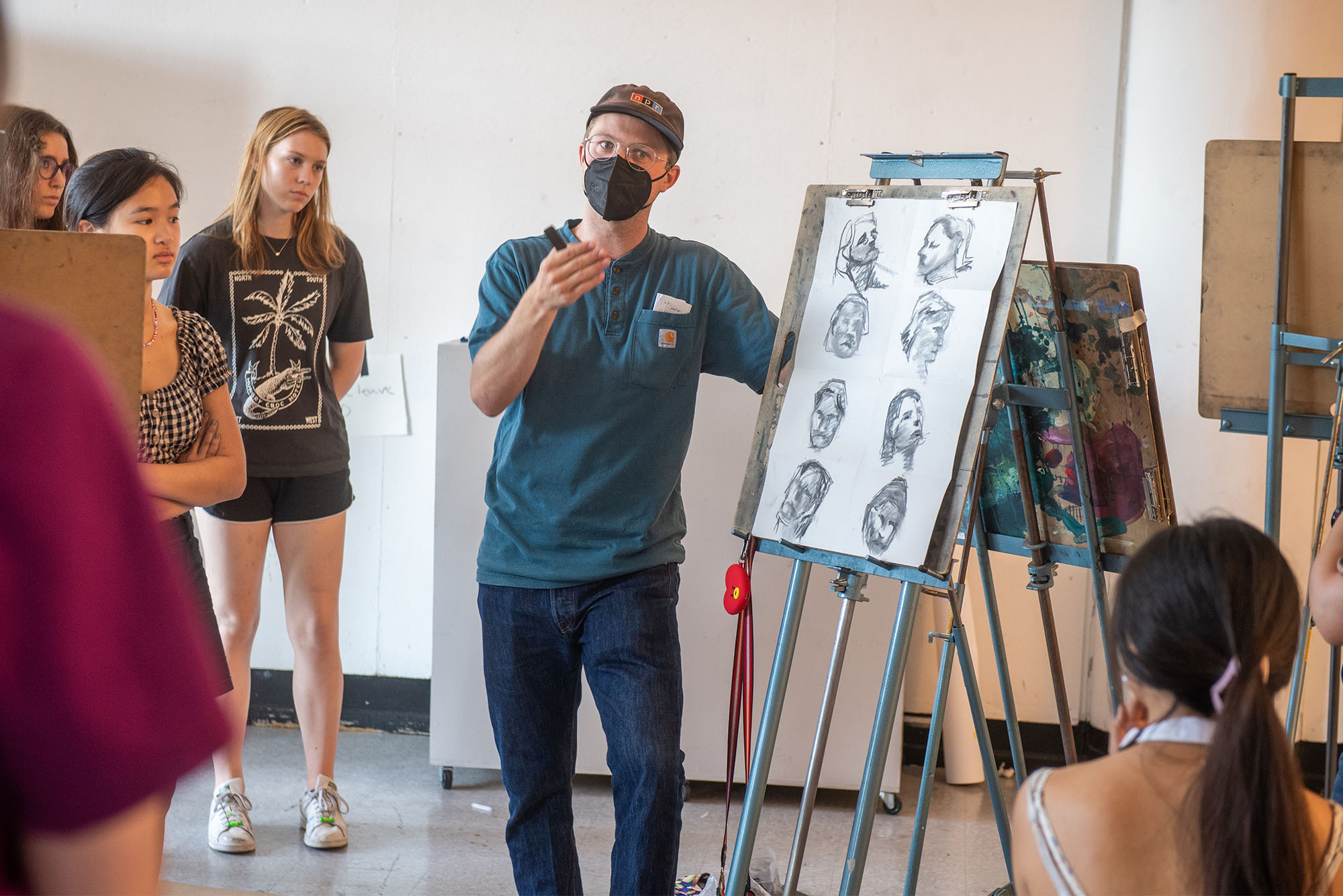 Photo of Instructor Sam Guy with his VASI drawing class. A masked man wearing a blue VASI labeled shirt and black face mask speaks to students as he stands in front of a metal easel featuring various charcoal sketches of heads. He holds a stick of charcoal in his hand as he gesticulates.