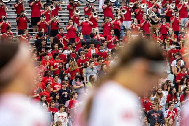 Photo of the Boston University Band wearing matching red shirts and black pants as they play their instruments in a middle stand. Inthe foreground are blurry profiles of two BU women soccer players in white uniforms.