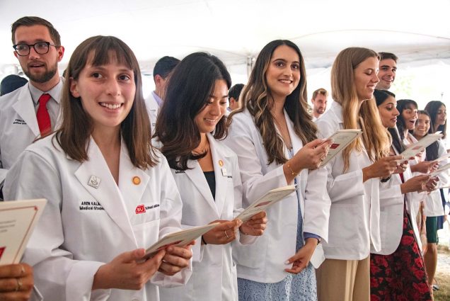 Photo of roughly six first-year medical students holding programs and wearing white coats with their names during the ceremony in which they recite the Hippocratic Oath for the first time, signaling their entrance to the study of medicine. The majority of the students in the row are women, White and AAPI, with a few men seen in the background. A white tent is seen behind them.
