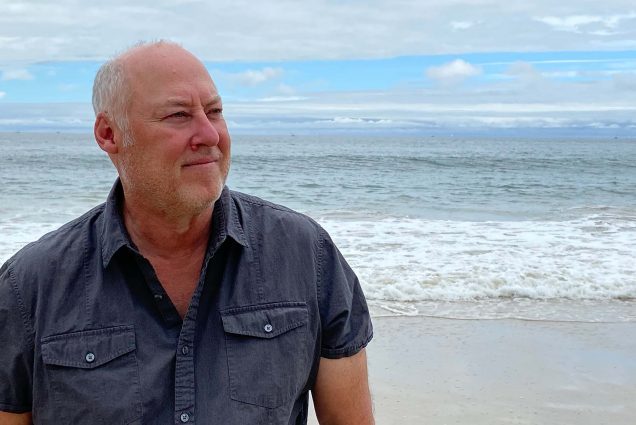 Photo of Stewart O’Nan, a middle age white man, who wears a dark gray short-sleeve button-down shirt and stands at the shore of a beach. He looks off to his left, and has short white hair and facial hair. The sky behind him is light blue with gray clouds.