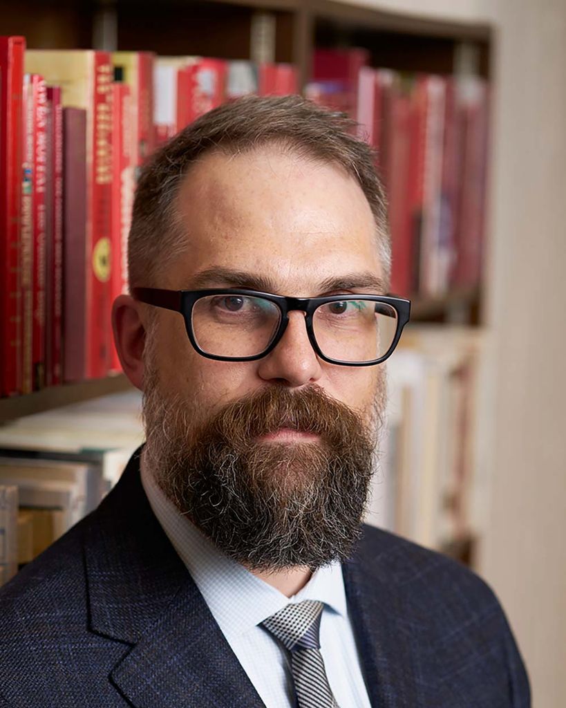 Photo of Mark Newton, a youngish White man with short brown hair, a long, brown beard and dark-rimmed glasses. He wears a suit and tie and stands in front of book case and makes a serious face. 