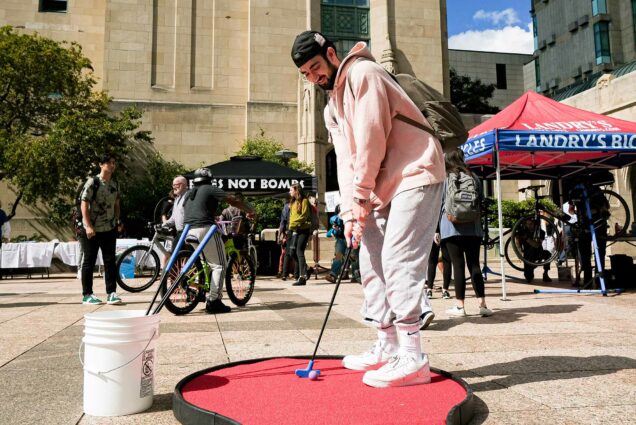 Photo of Tony Gluskin (COM'23) plays the mini golf at the Sustainability Festival on Marsh Plaza. A young man wearing a light pink hoodie, grey sweats, and black backpack puts on a small mini gold course. Sustainability stands featuring bike safety stands sit behind him.