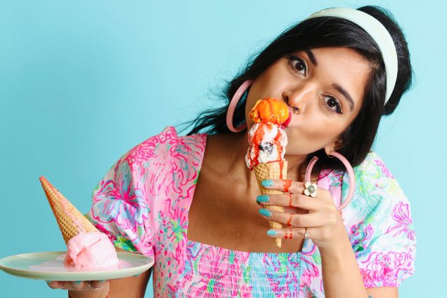 Photo of Chef Priyanka Naik, wearing a brightly colored floral dress, and holding a plate with an ice cream cone in her right hand, and eating an ice cream cone dripping with an orange sauce in her left hand. She wears a light blue headband, pink hoops, and stands in front of a bright blue background.