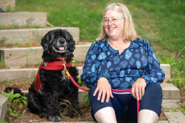 Photo: CGS professor Kari Lavalli, a white woman with shoulder-length blonder hair wearing a blue floral blouse and black pants, sits and poses with her dog, a black, seven-year-old Portuguese Water Dog Bell.