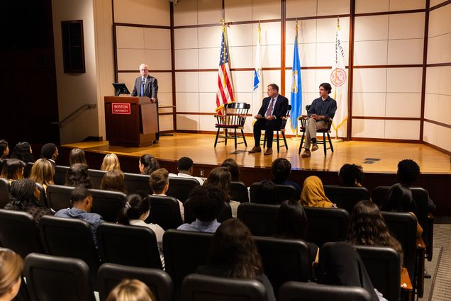 Photo of BU President Bob Brown giving a speech on stage. Two people are seated beside him on stage as he stands and address the crowd at a podium.