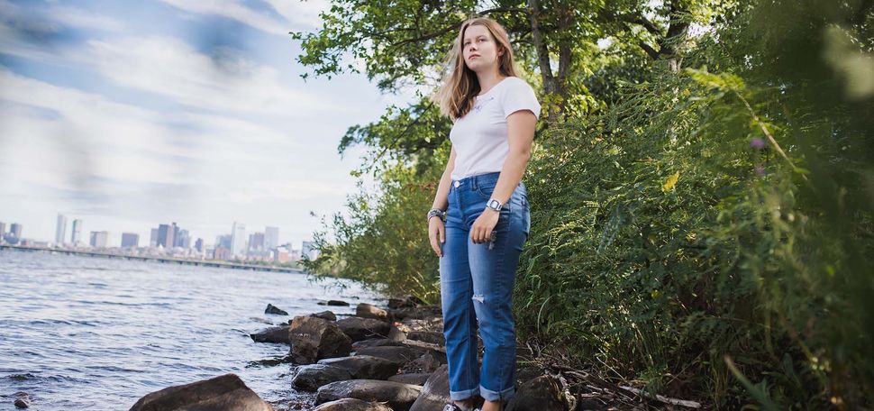 Photo: Natalia Sawicka, a young white woman with long blonde hair, poses for a photo on the bank of the Charles River. The city of Boston can be seen distantly in the background.