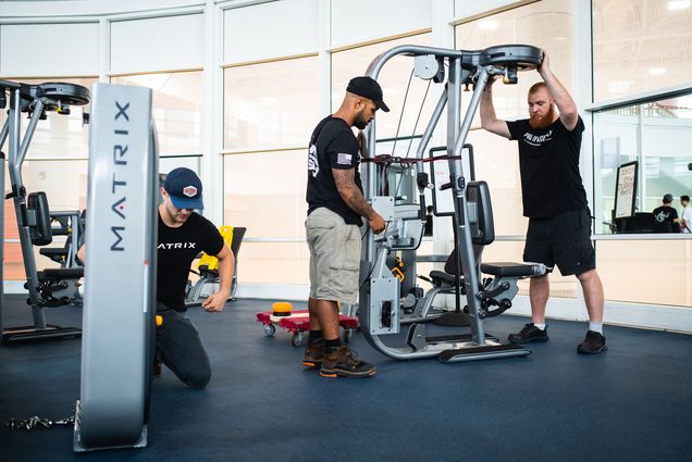 Photo of Jose Rodriquez (left) and Joe Moser of Pro Gym Supply setting up new equipment at Fit Rec. Two men wearing black shirts and shorts fiddle with large fitness equipment.