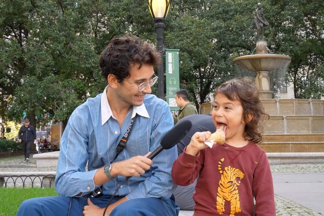 Image: screenshot of Julian Shapiro-Barnum, a young white man wearing glasses and a light blue sweater over a white collared shirt, interviewing a small child eating ice cream. Julian holds a black microphone to the child.