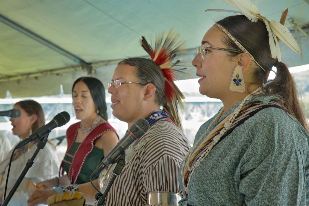 Photo of the Kingfisher Singers, a Wampanoag group singing. In the foreground, a woman with traditional beads, glasses, and a small feather hair piece is seen singing. Next, a man with a white and red quills in his pony tail is seen singing. Two other women are seen singing at the back left.