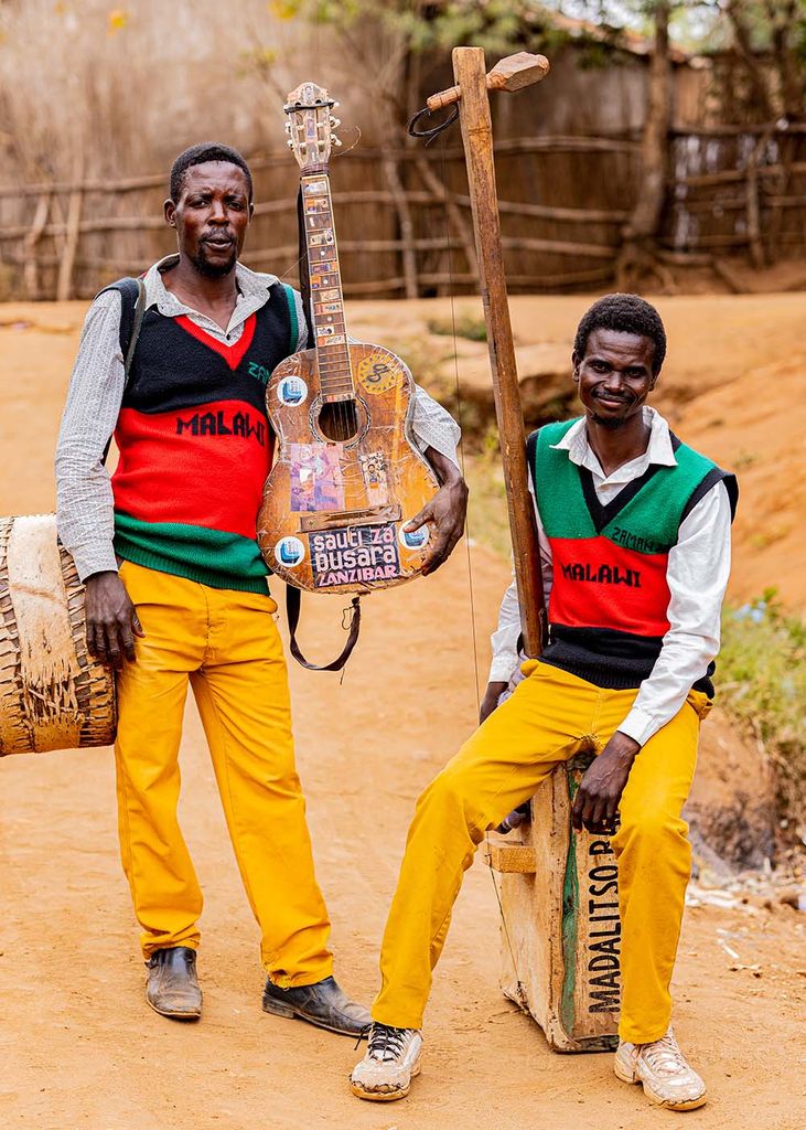 Photo of Madalitso, a pair of street musicians from Malawi, who pose with their acoustic instruments. At left, a youngish Black man holds a guitar in his left hand and has a drum at his hip; he wears a black, red and green Malawi sweater and yellow pants. At right, a youngish Black man wears the same, and sits atop an acoustic bass constructed from a wooden box and long beam.