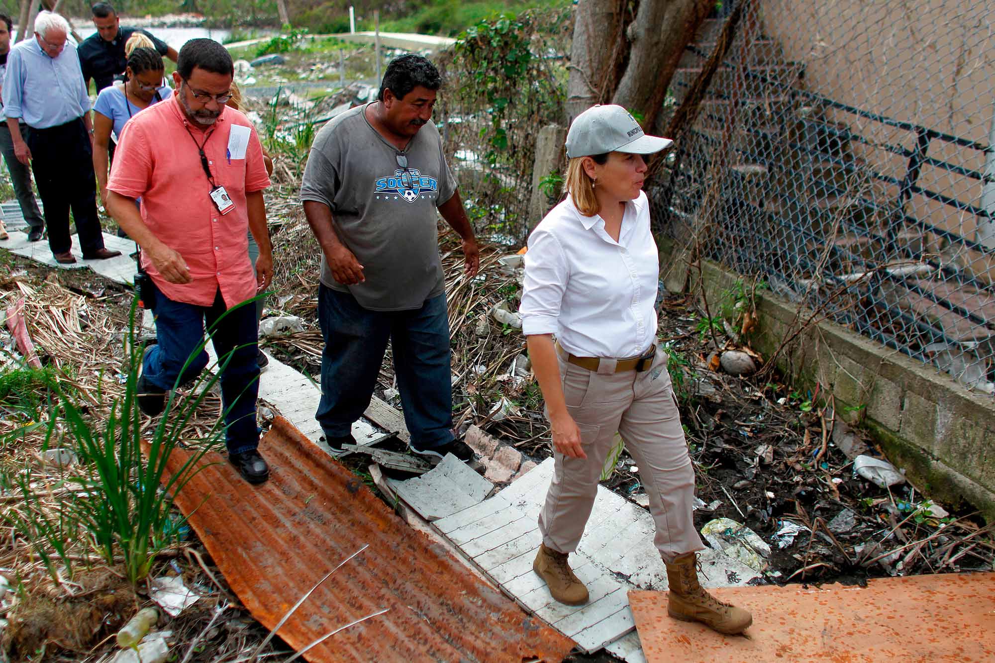 Photo of then mayor of San Juan, Carmen Yulín Cruz Soto touring debris from Hurricane Maria during a visit to the Playita community in San Juan, October 27, 2017. A woman wearing a cap, white shirt, and jeans leads a group of officials, including Bernie Sanders, through debris.