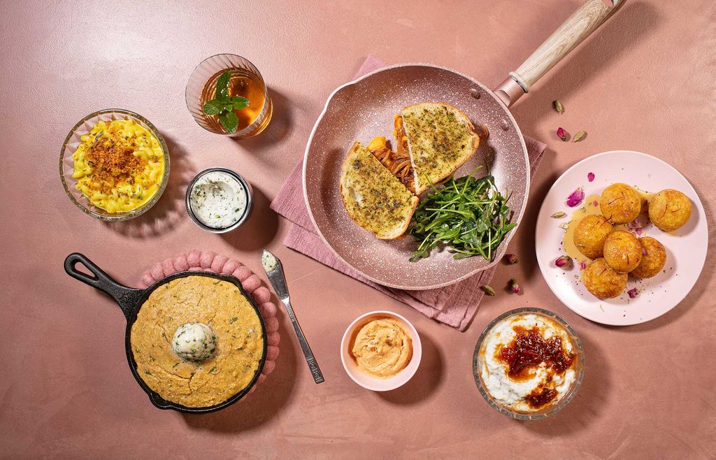 Aerial photo of the dishes included in the “The American Comfort Tiffin” chapter of Naik’s work. The dishes are laid on an almost sparkling, pale pink background, and include Chili-Maple Skillet Corn Bread (bottom left) and Masala Grilled Cheese (top middle) in a frying pan.