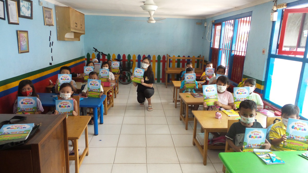 Photo of the class at the tiny shantytown helped by Manua’s Reading Train organization showing off the reading primer she wrote for the class. A masked young woman kneels in the middle of a classroom and holds up a small book. Various masked students seated at small desks in the classroom hold up copies of the same book.