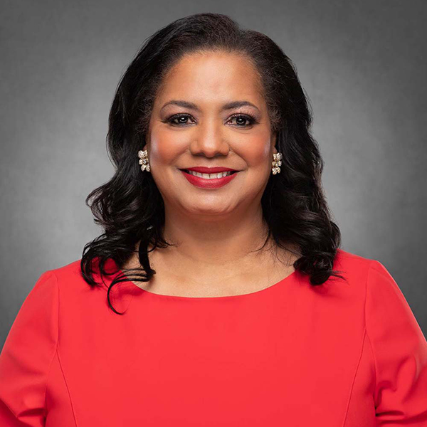 Photo of Antoinette “Tonie” Leatherberry (ENG’85), a middle-aged Black woman with shoulder length, slightly curled dark hair. She smiles, wears read lipstick, and earrings in the shape of a flower. She wears a pinkish-orange dress and stands in front of a dark gray background.