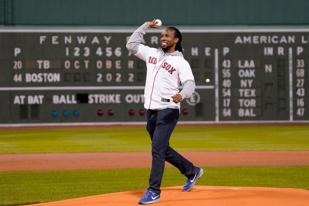 Photo: Dr. Ibram X. Kendi throws out the ceremonial first pitch before the start of a baseball game between the Boston Red Sox and the Tampa Bay Rays at Fenway Park in Boston. A Black man wearing a white and red Red Sox jersey top and jeans smiles awkwardly as he raises his arm up to throw a baseball.