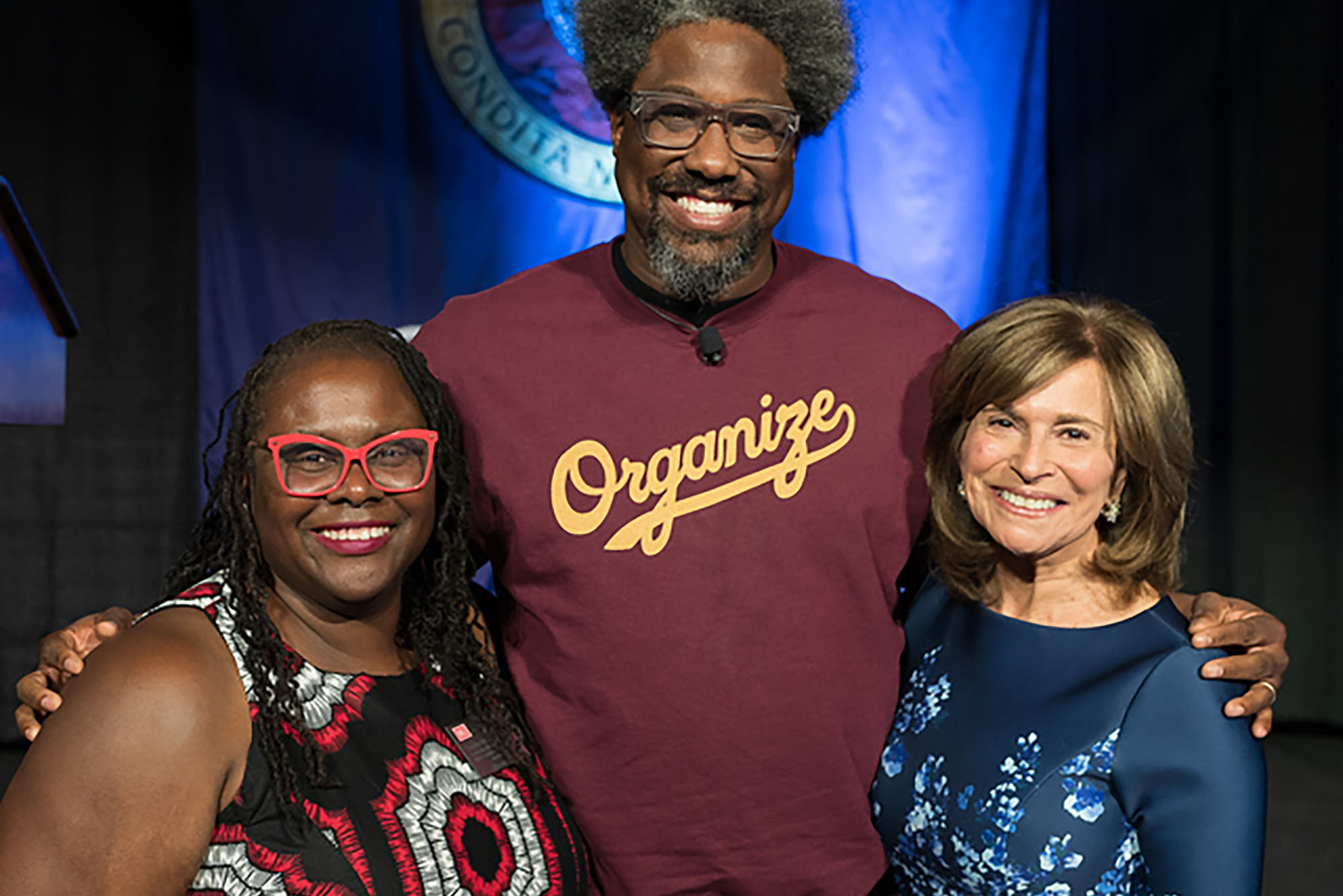 Photo: Dean Onwuachi-Willig (left) with W. Kamau Bell (center), and Rikki Klieman. A tall black man with graying bear and afro poses for a photo with arms over the shoulders of two women. On the left, a shorter black woman with long dreads, large red glasses, and a black and red dress smiles and on the right a shorter white woman with shoulder length brown hair and blue dress smiles.