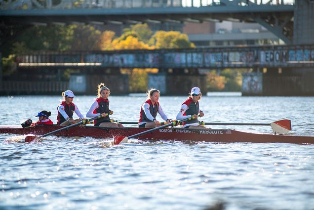 Photo: The BU women’s lightweight rowing team practicing on the Charles River ahead of this weekend’s Head of the Charles Regatta. Four young women wearing BU wet suits row down the Charles. A blurry bridge can be seen in the distance.