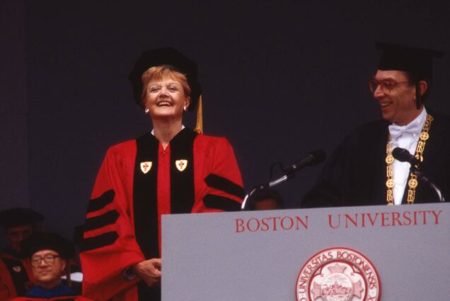 Photo: Angela Lansbury presented with an honorary degree by Jon Westling (Hon.’03), then BU president ad interim, at the University’s May 20,1990, Commencement. Lansbury stands on stage wearing a red ceremonial graduation gown as Westling stands behind a podium to her right in a black set of robes.