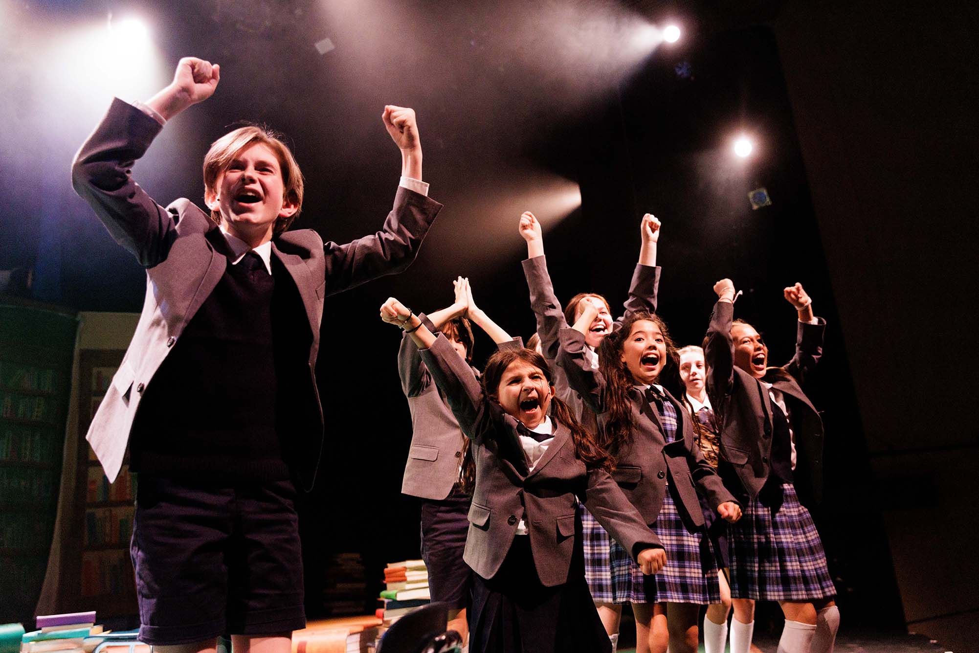 Photo: The youth ensemble performing the rousing number “Revolting Children,” during a dress rehearsal of "Roald Dahl’s Matilda, the Musical". A group of children wearing brown and black themed school uniforms all jump and cheer for joy, some with arms raised int he air.